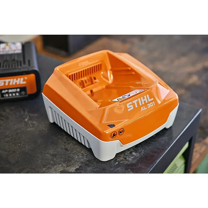 Stihl Akku-Schnellladegerät AL 301 - Passend für: STIHL Akkus AK, AP und AR - Nahaufnahme eines orangefarbenen Stihl AL 301 Ladegeräts mit weißer Basis auf einer dunklen Werkbank, mit einem Akku im Hintergrund.