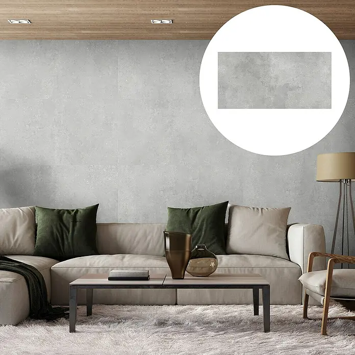 Revêt Wandpaneel SPC Wall - 1.200 x 600 mm, Beton Grau - Nahaufnahme der grauen Wandfliese mit Betonoptik. Die Fliese wird durch eine große, kreisförmige Lupe hervorgehoben, im Hintergrund ein Wohnzimmer.