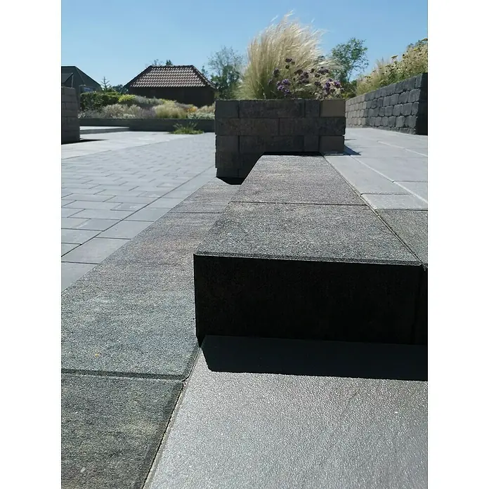 Diephaus Blockstufe Niles - 100 x 35 x 15 cm, Quarzit, Beton - Eine Nahaufnahme eines Pflastersystems mit grauen Steinen und einer bepflanzten Steinmauer im Hintergrund unter einem sonnigen Himmel.