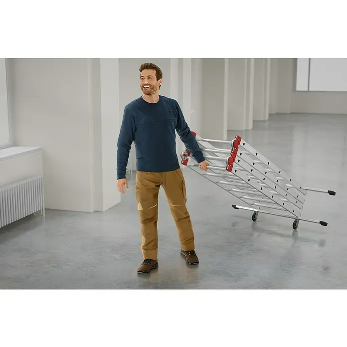 Hailo Leitergerüst G60 - Arbeitshöhe: 433 mm, Breite Plattform: 136 cm, Länge Plattform: 50 cm - Ein Mann trägt eine zusammengeklappte, fahrbare Mehrzweckleiter in einem hellen Raum. Die Leiter ist aus Aluminium gefertigt und hat rote Akzente.