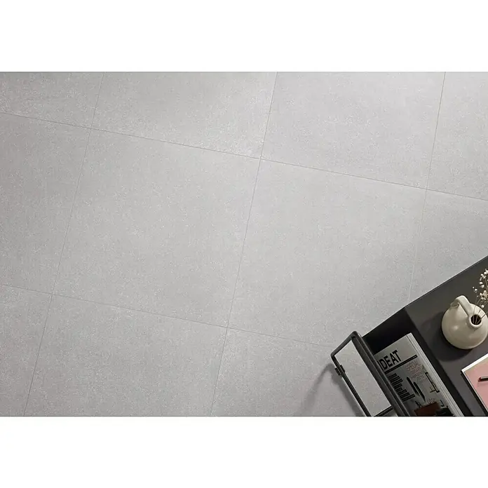 Pavimento porcelánico Weekend - 60 x 60 cm, Gris, Mate, Rectificado - Un revestimiento de suelo gris claro con un patrón de baldosas, parcialmente cubierto por una mesa de centro oscura con decoraciones.