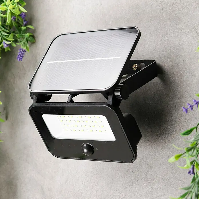 Arte confort Foco solar Bane - 1,5 W, L x An x Al: 14,3 x 5 x 10,2 cm, Negro, IP65, 400 lm - Primer plano de una luz solar negra de exterior montada en una pared, rodeada de vegetación.