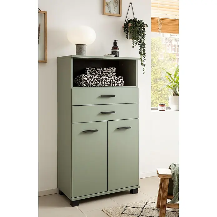 Schildmeyer Palermo Highboard - B x T x H: 60 x 32,6 x 110,5 cm, Pistazie, Matt - Ein grüner Schrank mit Schubladen und Türen steht in einem hellen Raum, darauf eine Lampe und Handtücher.