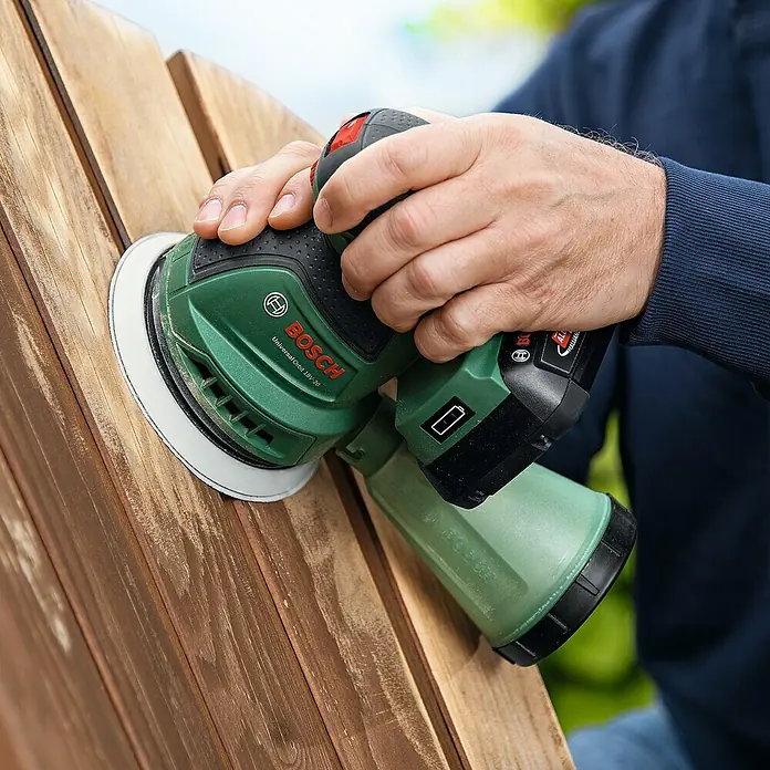 Bosch Power for All 18V Akku-Exzenterschleifer UniversalOrbit 18V-20 - 18 V, Ohne Akku, Durchmesser Schleifteller: 125 mm - Nahaufnahme eines Mannes, der mit einem grünen Bosch-Schleifgerät Holz schleift, wobei der Fokus auf dem Werkzeug und dem Schleifprozess liegt.