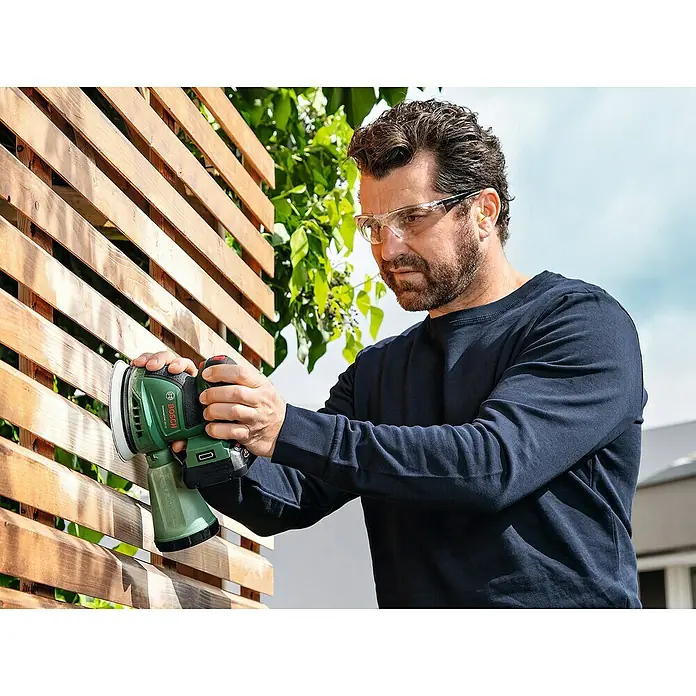 Bosch Power for All 18V Akku-Exzenterschleifer UniversalOrbit 18V-20 - 18 V, Ohne Akku, Durchmesser Schleifteller: 125 mm - Ein Mann schleift mit einem Bosch Exzenterschleifer ein Holzgitter im Freien und trägt dabei eine Schutzbrille.