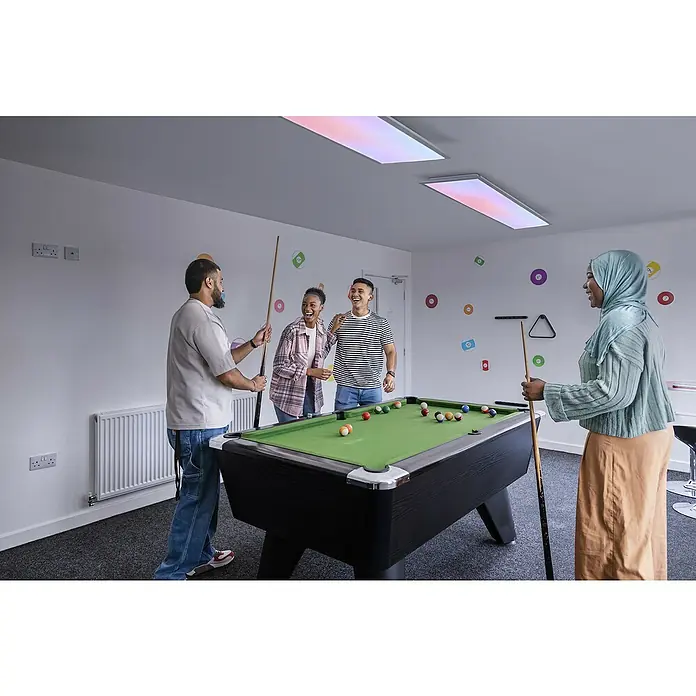 Osram Smart+ LED-Panel Planon Magic RGB + Tunable White - 36 W, L x B x H: 119,5 x 29,5 x 6 cm, Weiß, RGBW - Gruppe von Freunden spielt Billard auf einem schwarzen Billardtisch mit grünem Tuch, umgeben von Dekorationen.