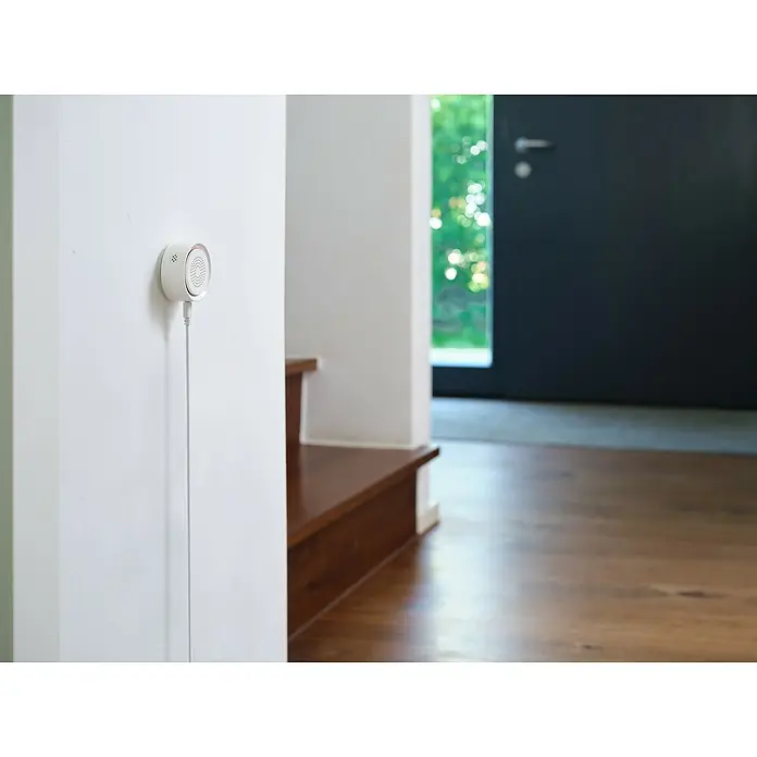 Osram Smart+ Alarmsirene - Innen, 90 dB, Netzbetrieben - Ein weißer, runder Luftqualitätsmonitor ist an einer weißen Wand befestigt und mit einem Kabel verbunden.