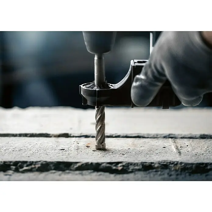 Bosch Beton- & Steinbohrer PRO SDS plus-5X - 1 Stk., Durchmesser Bohrer: 12 mm, Arbeitslänge: 100 mm - Nahaufnahme einer Bohrmaschine mit Betonbohrer, die ein Loch in eine graue Betonfläche bohrt. Eine Hand mit Handschuh ist zu sehen.