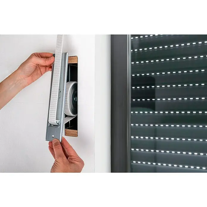Schellenberg Einlasswickler - 244 x 50 x 140 mm, Lochabstand: 215 mm, Geeignet für: Rollladen-Maxi-Systeme - Nahaufnahme eines Rollladenkastens mit Gurtwickler, der in eine weiße Wand eingebaut ist und von Händen gehalten wird.