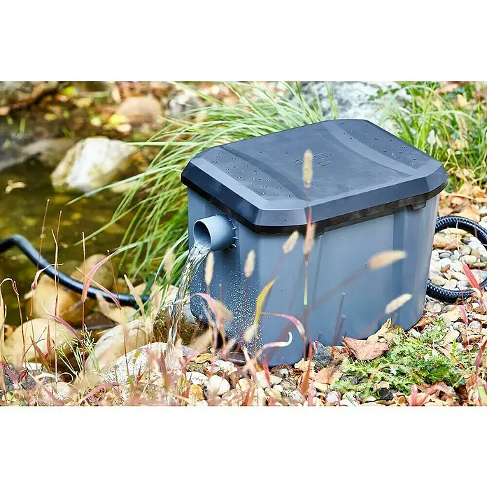 Heissner Durchlauffilter-Set - 33 W, Teichgröße: 8.000 l - Nahaufnahme eines Heissner Teichfilters im Freien mit Wasser, das aus einem Auslass fließt, umgeben von Steinen und Pflanzen.