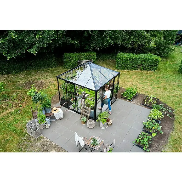 Halls Gewächshaus Qube Square - Außenmaß inkl. Dachüberstand (B x T): 250,8 x 250,8 cm, Material Seitenwand: Glas, 3 mm, Schwarz - Ein Gewächshaus mit Glaswänden und schwarzem Rahmen steht im Garten, umgeben von Pflanzen, Töpfen und Gartenmöbeln für entspannte Stunden.