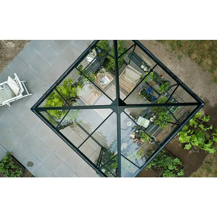 Halls Gewächshaus Qube Square - Außenmaß inkl. Dachüberstand (B x T): 250,8 x 250,8 cm, Material Seitenwand: Glas, 3 mm, Schwarz - Luftaufnahme eines Gewächshauses mit Pflanzen, Sitzbereich und Steinboden. Das Gewächshaus bietet einen geschützten Raum für Pflanzenliebhaber.