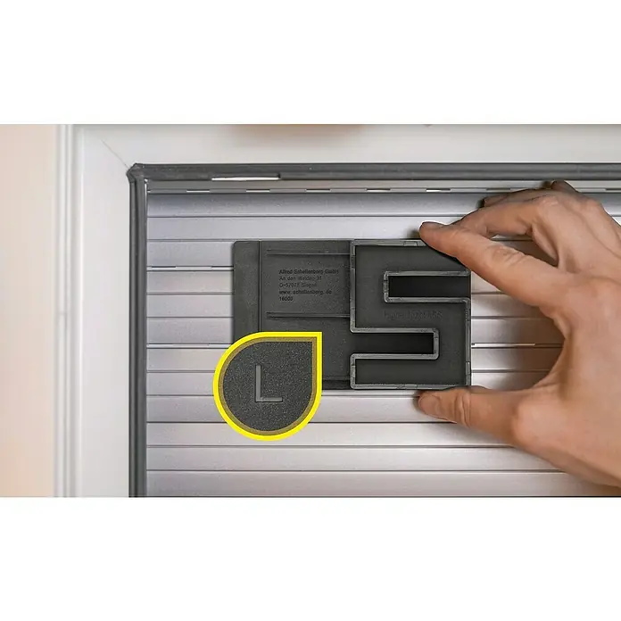 Schellenberg Hochschiebesicherung - 2 Stk. - Nahaufnahme eines Rollladenkastens mit einer schwarzen Kunststoffform, die die Buchstaben "S" und "L" enthält, zur Rollladenreparatur.