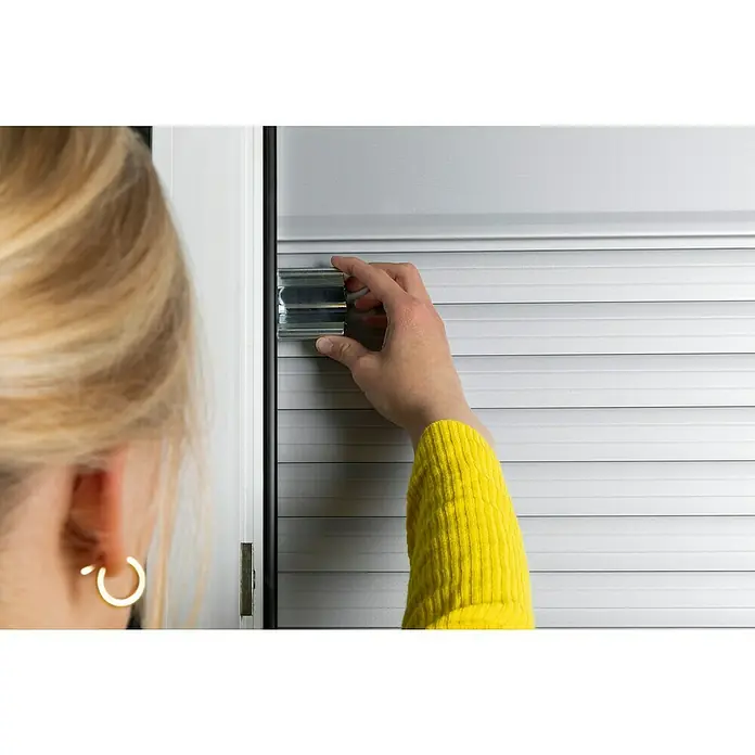 Schellenberg Klemmsicherung - Geeignet für Profil-Deckhöhe: Ab 45 mm, 2 Stk. - Eine Person befestigt eine silberne Jalousie an einem Fensterrahmen, wobei der Fokus auf der Jalousie und der Befestigung liegt.