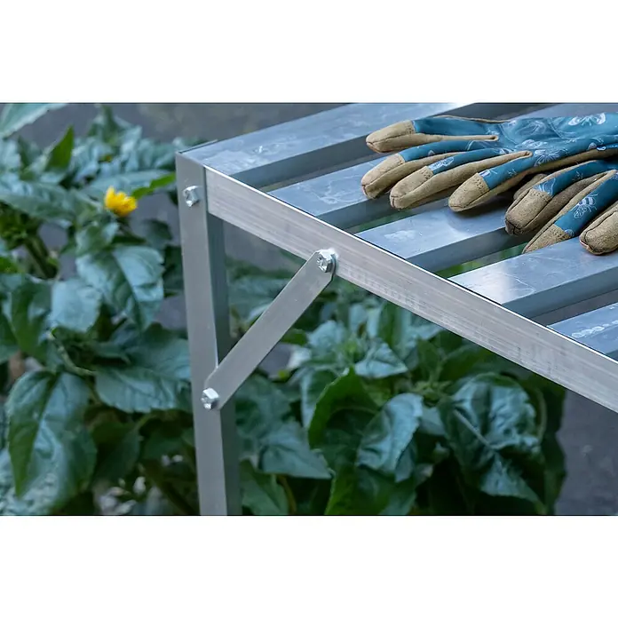 Halls Gewächshaus-Tisch - 120,8 x 76 cm, Aluminium, Eine Ablageebene - Nahaufnahme eines verzinkten Gartentisches mit Arbeitshandschuhen auf der Oberfläche, umgeben von üppigem Grün.