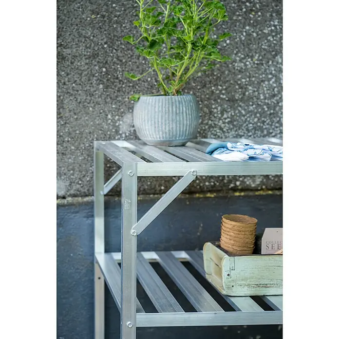 Halls Gewächshaus-Tisch - 120,8 x 76 cm, Aluminium, Doppelte Ablageebene - Nahaufnahme eines silbernen Pflanztisches mit einer Topfpflanze, Gartenhandschuhen und Gartenzubehör vor einer grauen Wand.