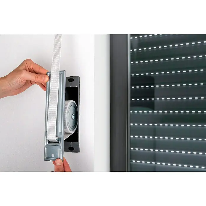 Schellenberg Rollladen-Gurtwickler Maxi - Lochabstand: 185 mm, Gurtbreite: 23 mm, Max. Gurtlänge: 5,6 m - Nahaufnahme einer Person, die einen Rollladengurtwickler an einer weißen Wand montiert, neben einem grauen Rollladen für Fenster.