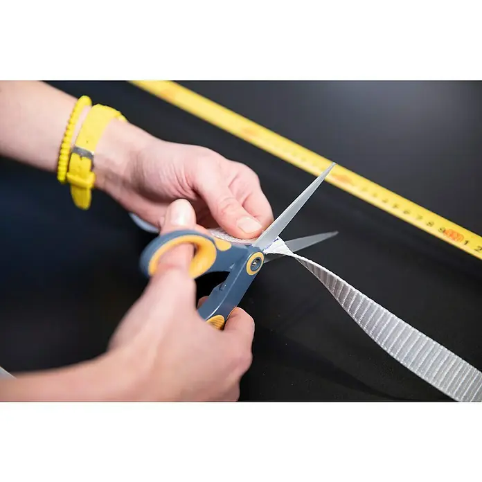 Schellenberg Rollladengurt Maxi - Grau, Länge: 6 m, Gurtbreite: 23 mm - Eine Person schneidet ein weißes Textilband mit einer Schere, ein gelbes Maßband liegt daneben auf einem schwarzen Tisch.