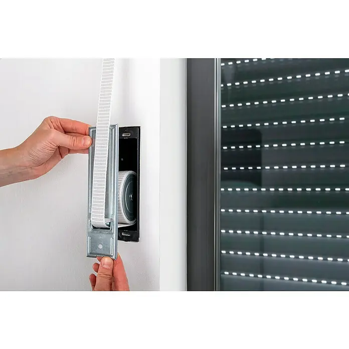 Schellenberg Rollladen-Gurtwickler Maxi - Lochabstand: 160 mm, Gurtbreite: 23 mm, Max. Gurtlänge: 5,3 m - Nahaufnahme einer Person, die einen Rollladen-Gurtwickler an einer Wand montiert, wobei der Fokus auf dem Gurt und der Mechanik liegt.