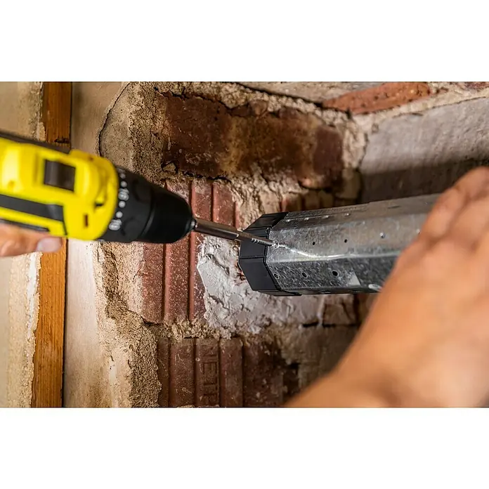 Schellenberg Walzenhülse Mini - Durchmesser Achtkantwelle: 40 mm, Länge: 104 mm, Geeignet für: Rollladen-Mini-Systeme - Ein Handwerker befestigt mit einem gelben Akkuschrauber ein silbernes Metallteil an einer Ziegelwand, wobei Schrauben verwendet werden.