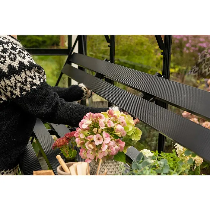 Halls Gewächshaus-Regal - B x L: 2,5 x 235 cm, Schwarz, Passend für: Halls Gewächshaus QUBE 8 mit 6,3 m² - Nahaufnahme eines schwarzen Gewächshausregals mit Blumen und einer Person im Hintergrund.