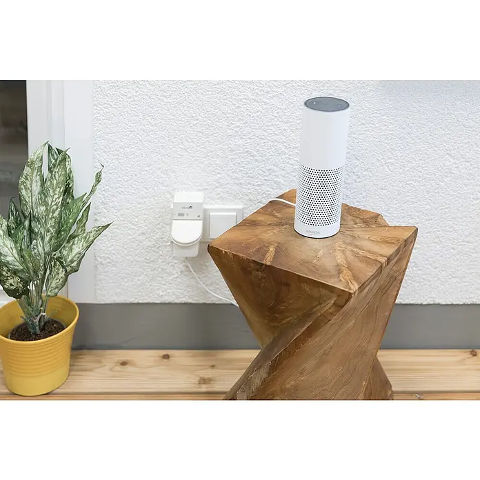 Schellenberg Smart Home Funksteckdose - Außen, Weiß - Der Amazon Echo Lautsprecher steht auf einem Holzhocker neben einer Grünpflanze, ein smartes und modernes Zuhause.