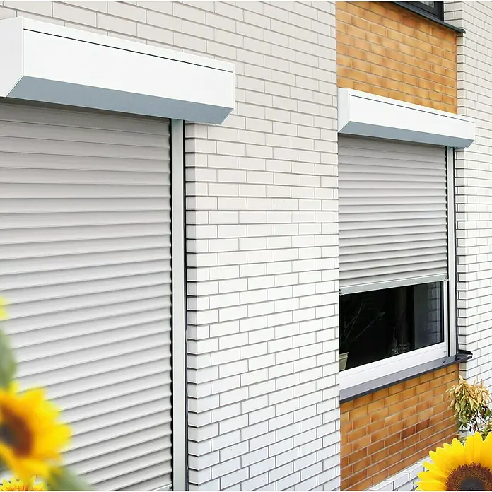 Schellenberg Rollladen-Komplett-Set MINI - Vorbau, 80 x 100 cm - Zwei Fenster mit Rollläden an einer Ziegelwand, eines teilweise geöffnet. Die Rollläden sind in hellgrauer Farbe gehalten und bieten Sonnenschutz.
