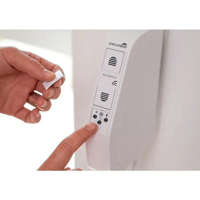 Schellenberg Funk-Rollladen-Gurtwickler Rollo Drive 75 Premium - L x B x H: 155 x 37 x 199 mm, Gurtbreite: 14 mm - 15 mm, Zugkraft: 30 kg, Unterstützte Systeme: Schellenberg Smart Home System - Nahaufnahme eines Schellenberg RolloDrive 75 Gerätes mit einer Hand, die einen Knopf drückt und eine andere, die einen weißen Stecker hält.
