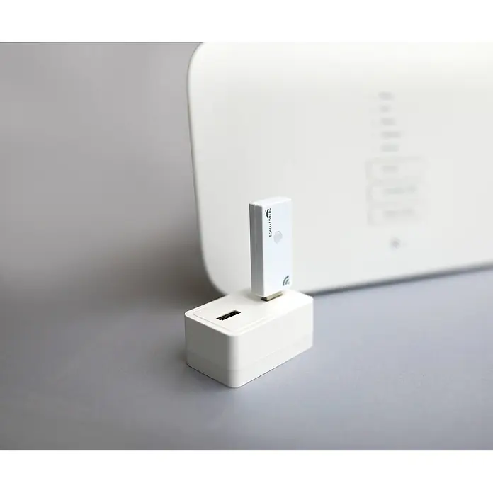 Schellenberg Smart Home Funksender Funk-Stick - Weiß, Reichweite ca.: 20 m - 100 m - Drahtloser Schellenberg-USB-Stick, der in einen Adapter gesteckt ist, und ein weißer Router dahinter.