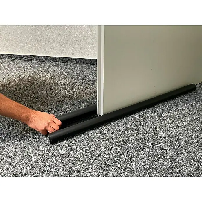Schellenberg Türbodendichtung Plus - Anthrazit, L x B: 95 cm x 35 mm, Geeignet für: Zimmertüren - Bild einer schwarzen, doppelten Zugluftstopper unter einer Tür, die von einer Hand angefasst wird, um sie zu justieren.