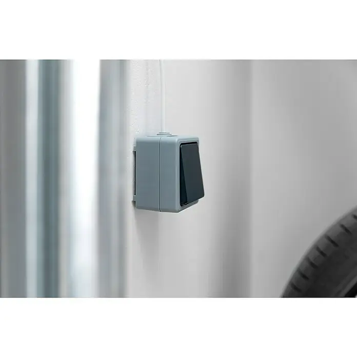 Schellenberg Innentaster - Passend für: Schellenberg Garagentorantriebe, Aufputzmontage - Nahaufnahme eines grauen Lichtschalters an einer Wand mit sichtbarem Kabel und Teil eines Fahrradreifens.