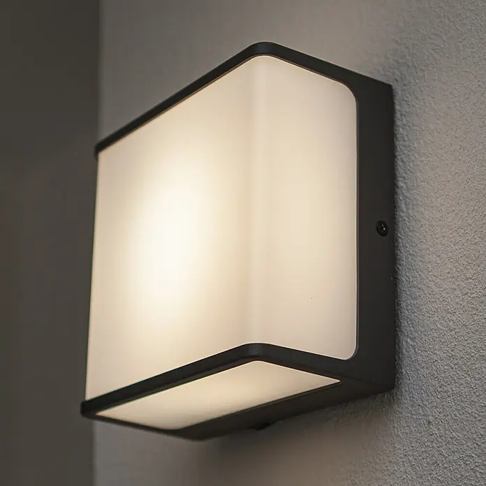 Lutec Doblo LED-Außenwandleuchte DOBLO - 9 W, 8 x 15 x 15 cm, Schwarz-matt, IP54 - Nahaufnahme einer quadratischen LED-Wandleuchte mit schwarzem Rahmen an einer grauen Wand, die ein warmes, diffuses Licht ausstrahlt.