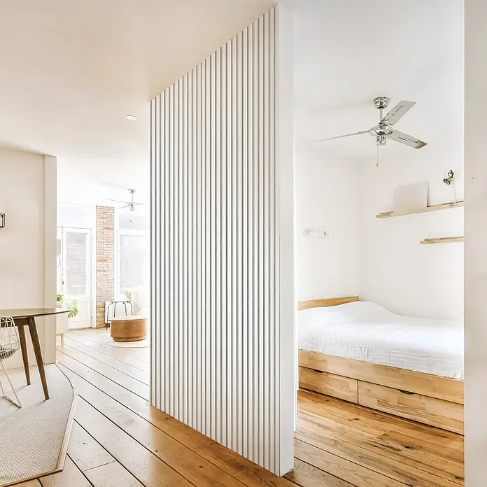 FibroTech Akustikpaneel Unika 25 - White Platinum, 2.440 x 600 x 22 mm - Ein minimalistisches Interieur mit einer weißen Lamellenwand, die Wohn- und Schlafbereich trennt, akzentuiert durch Holzböden und natürliche Elemente.