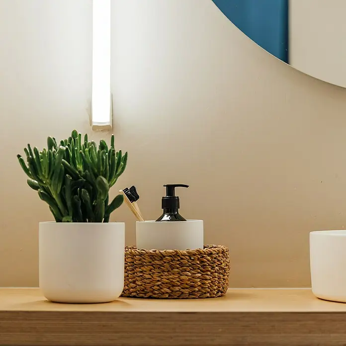 Maceta para planta Sita - Dimensiones externas (Ø x Al): 9,5 x 9,5 cm, Cerámica, Blanco - Arreglo de baño con una suculenta en una maceta blanca, un dispensador de jabón y cepillos de dientes de bambú en una cesta de mimbre sobre una estantería de madera.