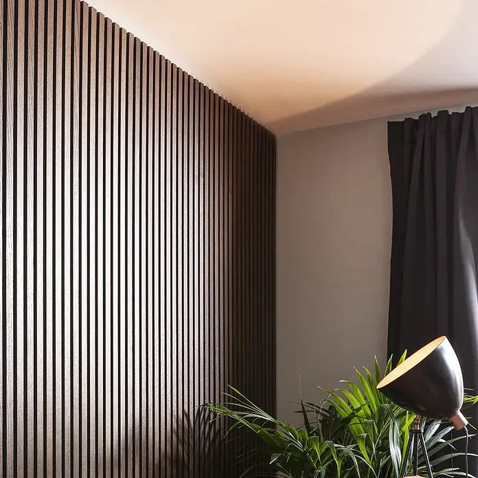 FibroTech Akustikpaneel Premium - Smoked Oak, 2.440 x 605 x 22 mm - Dieses Bild zeigt eine Akustikpaneelwand mit Holzlatten, die eine moderne und stilvolle Raumgestaltung ermöglicht. Im Vordergrund befindet sich eine Zimmerpflanze.
