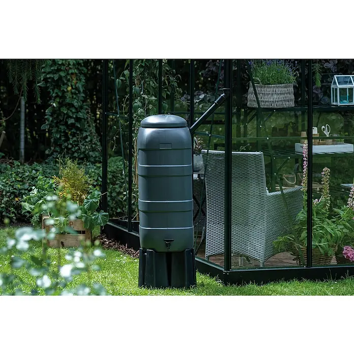 Halls Regentonne - Schwarz - Ein grauer Regenwasserbehälter mit schwarzem Sockel steht vor einem Gewächshaus in einem Garten.