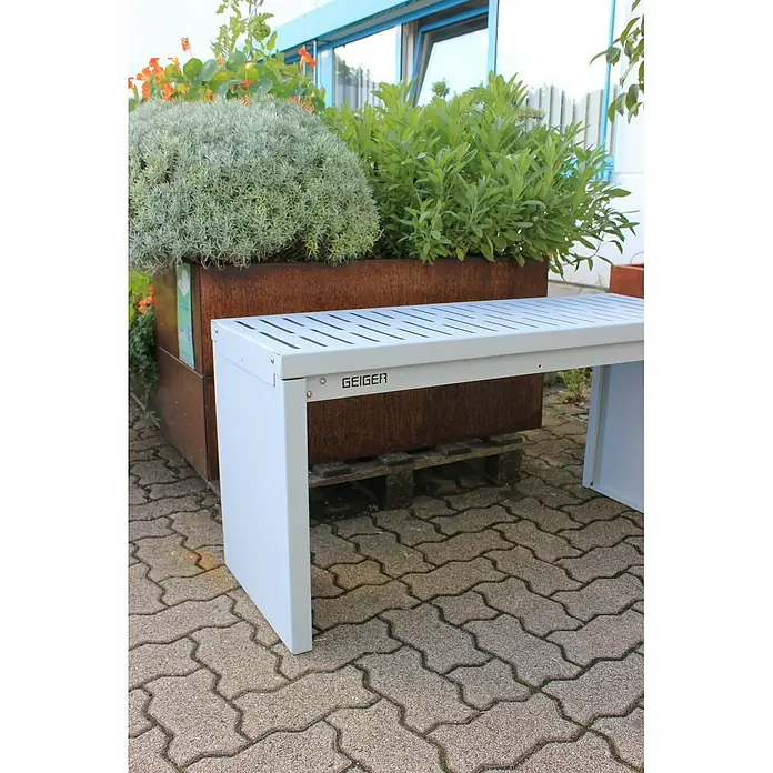 Palatino Bankauflage Bridge - L x B x H: 109 x 40 x 4 cm, Weiß - Eine moderne, weisse Bank der Marke Geiger steht neben einem Pflanzkasten mit üppigem Grün auf einem Pflaster.