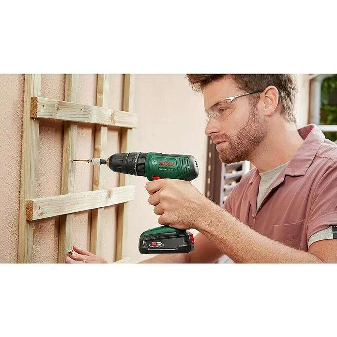 Bosch Power for All 18V Akku-Schlagbohrschrauber EasyImpact 18V-38 - 18 V, 1 Akku, 2,5 Ah, 38 Nm - Ein Mann befestigt mit einem Bosch Akku-Bohrschrauber ein Holzgitter an einer Wand, wobei er eine Schutzbrille trägt.