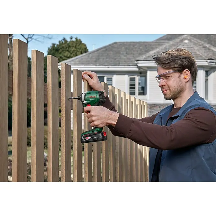 Bosch Power for All 18V Akku-Drehschlagschrauber UniversalImpactDrive 18V-210 - 18 V, Ohne Akku, 210 Nm - Ein Mann montiert einen Holzzaun mit einem grünen Bosch Akku-Bohrschrauber, der über einen 2.0 Ah Akku verfügt.