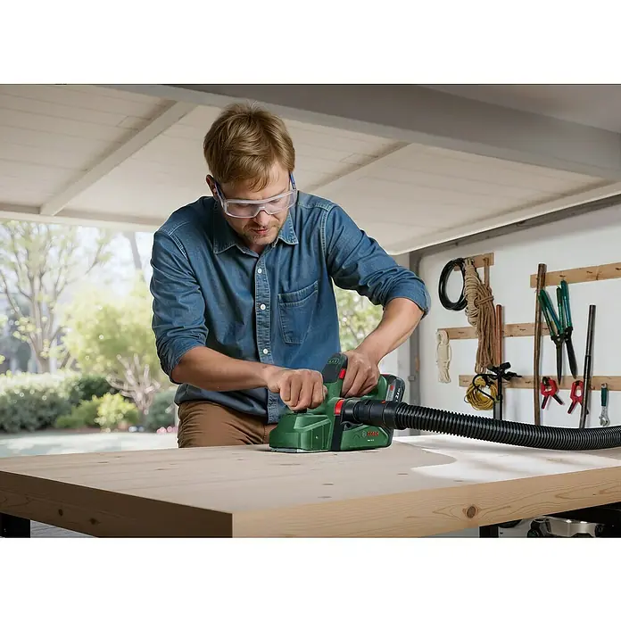 Bosch Power for All 18V Akku-Handhobel PHO 18V-20 - 18 V, Ohne Akku, Hobelbreite: 82 mm - Ein Mann verwendet einen grünen Bosch Hobel mit Staubabsaugung auf einem Holztisch in einer Werkstatt.