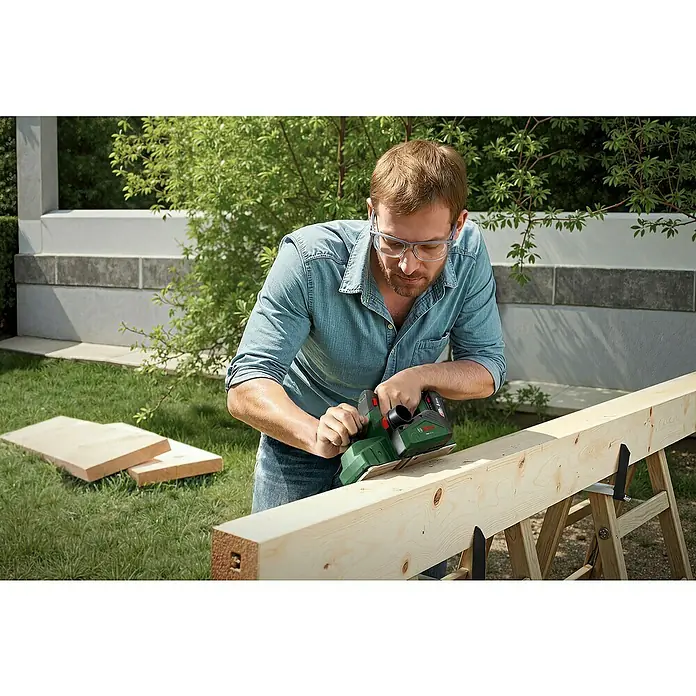 Bosch Power for All 18V Akku-Handhobel PHO 18V-20 - 18 V, Ohne Akku, Hobelbreite: 82 mm - Ein Mann hobelt ein Holzbrett im Garten mit einem grünen Bosch Elektrohobel, Schutzbrille tragend.