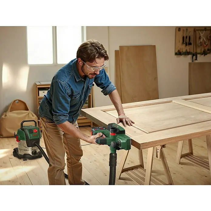 Bosch Power for All 18V Akku-Handhobel PHO 18V-20 - 18 V, Ohne Akku, Hobelbreite: 82 mm - Ein Mann benutzt einen Bosch Elektrohobel, um ein Holzstück zu bearbeiten, mit einem Staubsauger im Hintergrund.