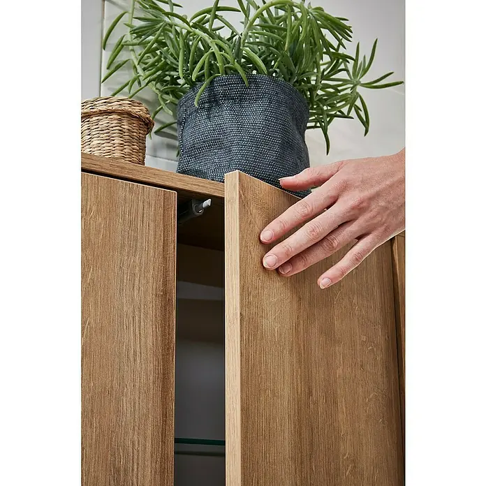 Fackelmann Finn Bad-Midischrank - B x T x H: 600 x 305 x 750 mm, Ast-Eiche - Nahaufnahme eines Holzschranks mit einer Hand, die ihn öffnet, und einer Pflanze darauf.