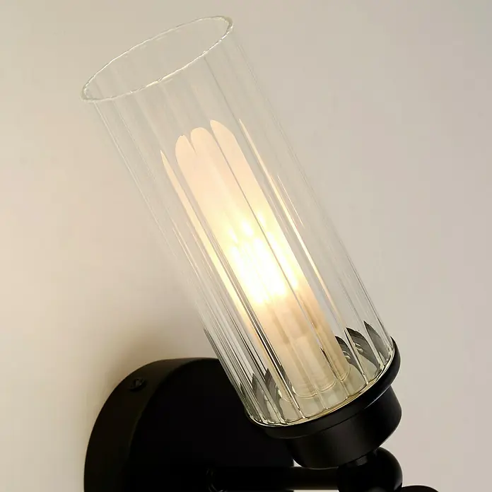 Searchlight Wandleuchte Imperial - 7 W, L x B x H: 37 x 14 x 10 cm, Schwarz, G9 - Nahaufnahme einer eleganten Wandleuchte mit einem klaren, geriffelten Glaszylinder, der ein warmes Licht ausstrahlt und eine stilvolle Atmosphäre schafft.