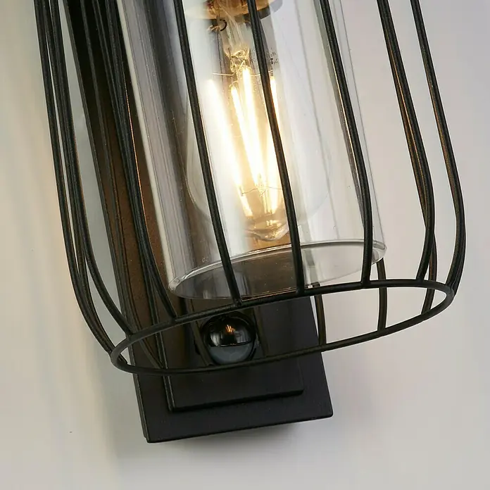 Searchlight Wandleuchte Reed - 40 W, L x B x H: 16 x 16 x 27,2 cm, Schwarz - Nahaufnahme einer industriellen Wandleuchte mit schwarzem Metallkäfig und Edison-Glühbirne, die ein warmes Licht ausstrahlt.
