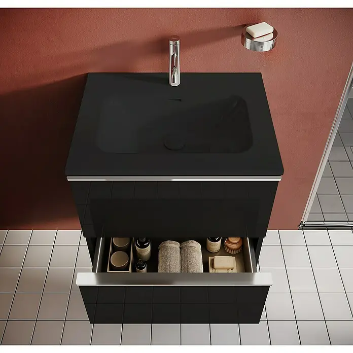 Riva Moon Waschtischunterschrank - B x T x H: 60 x 45,3 x 63,5 cm, Schwarz - Schwarzer Waschtisch mit offenem Schubfach, das Badutensilien wie Handtücher und Seife zeigt, an einer roten Wand.