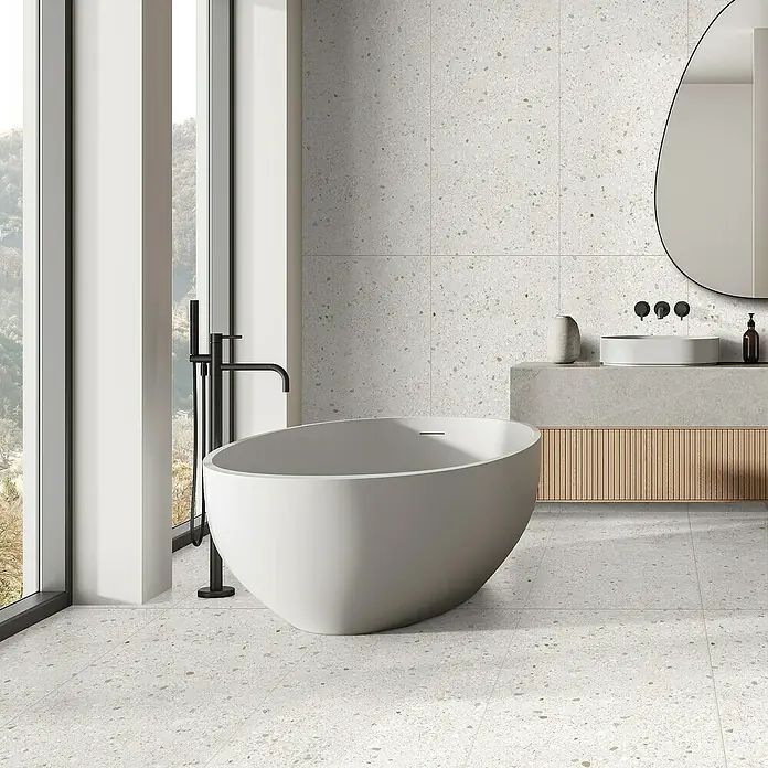 Feinsteinzeugfliese Mineral Stone - 60 x 119,8 cm, White, Matt - Elegantes Badezimmer mit freistehender Badewanne, Waschtisch und Spiegel vor einer stilvollen Terrazzo-Wand.
