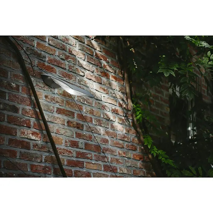 Lutec Aplique solar para exterior Zerta - 3 W, L x An x Al: 25,5 x 20 x 14,5 cm, Gris, IP44 - Una moderna luz de pared metálica ilumina una pared de ladrillo, rodeada de plantas. Proporciona iluminación exterior elegante.