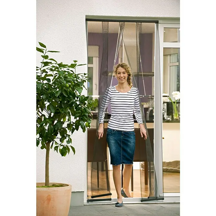 Tesa Insect Stop Mosquitera Standard - An x Al: 95 x 220 cm, Montaje de la mosquitera: Enganche, Antracita - Mujer cruzando una puerta mosquitera magnética. Ella está usando una blusa a rayas y una falda de mezclilla.