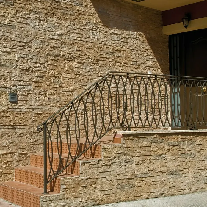 Revestimiento de piedra Salonica Canela - 60 x 30 x 2 cm - Foto detallada de una escalera exterior con una barandilla adornada frente a una fachada hecha de paneles de piedra decorativos.
